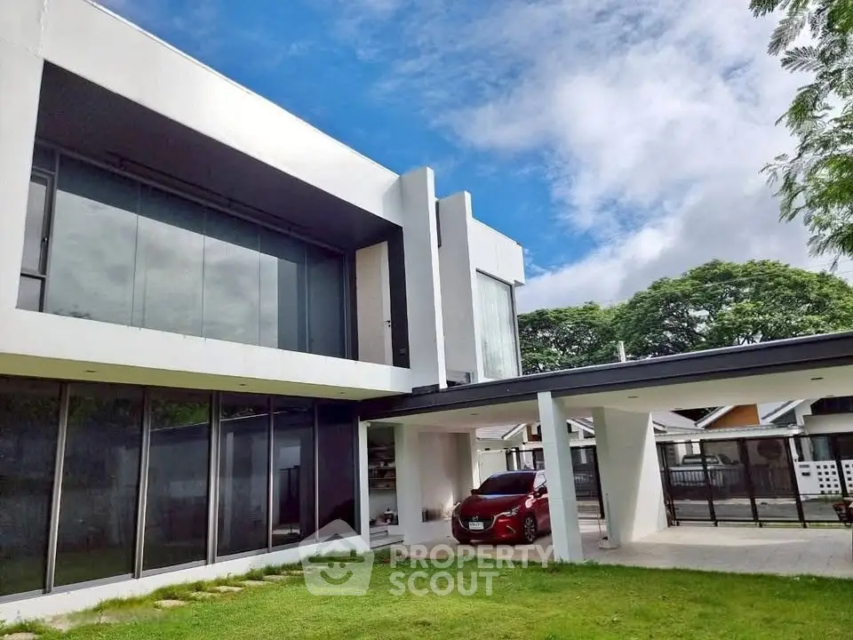 Modern two-story house with sleek design and spacious carport, surrounded by lush greenery.