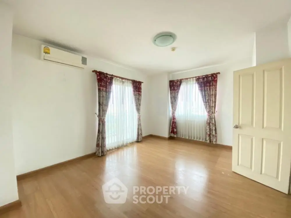 Spacious and well-lit living room with polished wooden flooring, large windows draped with elegant curtains, and a modern air conditioning unit.