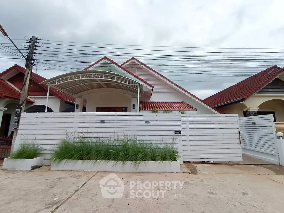 Charming single-story house with red roof and white fence, featuring a covered entrance and lush greenery.