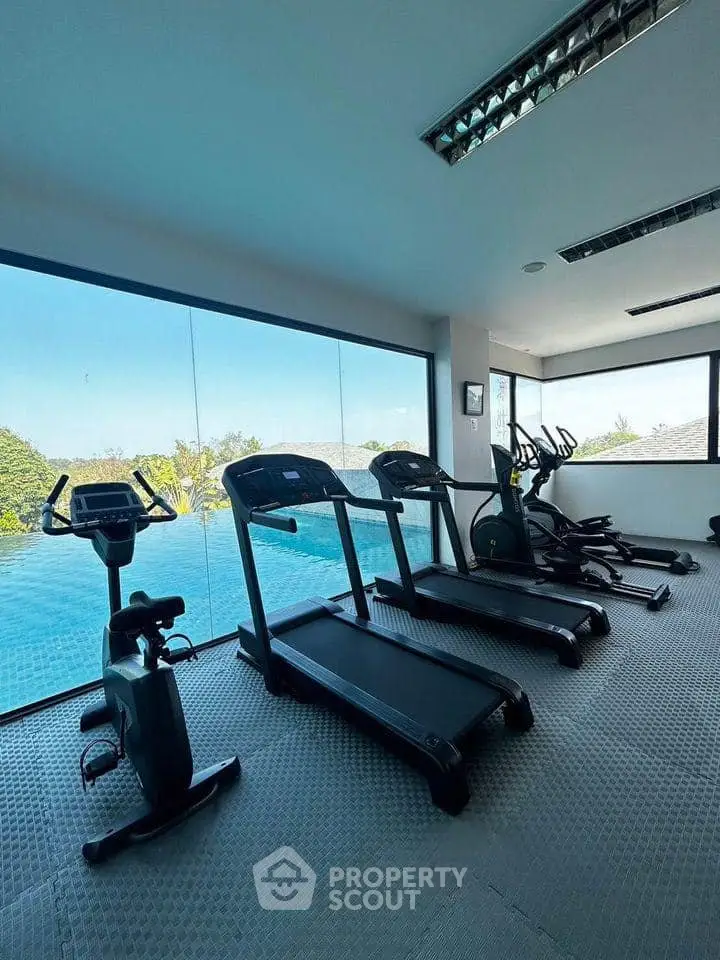 Modern gym with large windows overlooking a pool and lush greenery, featuring state-of-the-art exercise equipment.