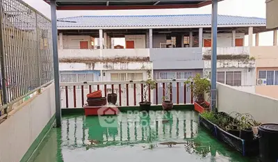 Spacious balcony with potted plants and covered roof, overlooking neighboring buildings.