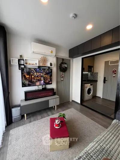 Modern living room with stylish decor, featuring a wall-mounted TV, cozy furniture, and a glimpse of a kitchen with washing machine.