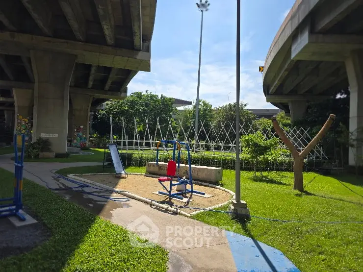Urban park under highway overpass with playground equipment and green landscaping.