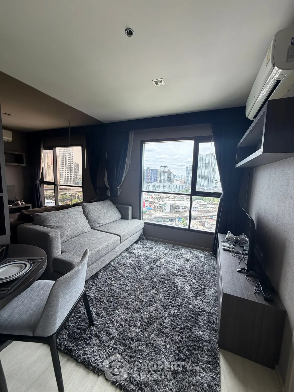 Modern living room with city view, featuring a cozy gray sofa, plush rug, and large windows.