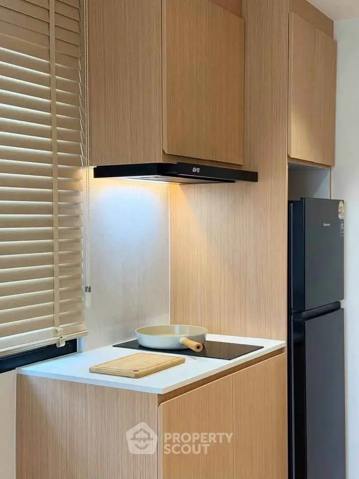 Modern kitchen with sleek wooden cabinets and built-in stovetop, featuring a compact fridge and window blinds.