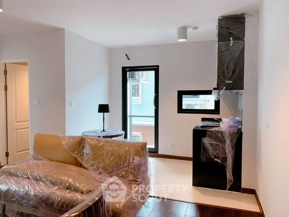 Modern living room with wrapped furniture and kitchen area, featuring sleek design and natural light.