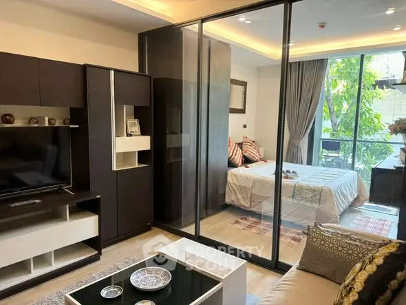 Modern open-plan living room with glass partition leading to a cozy bedroom, featuring stylish decor and natural light.