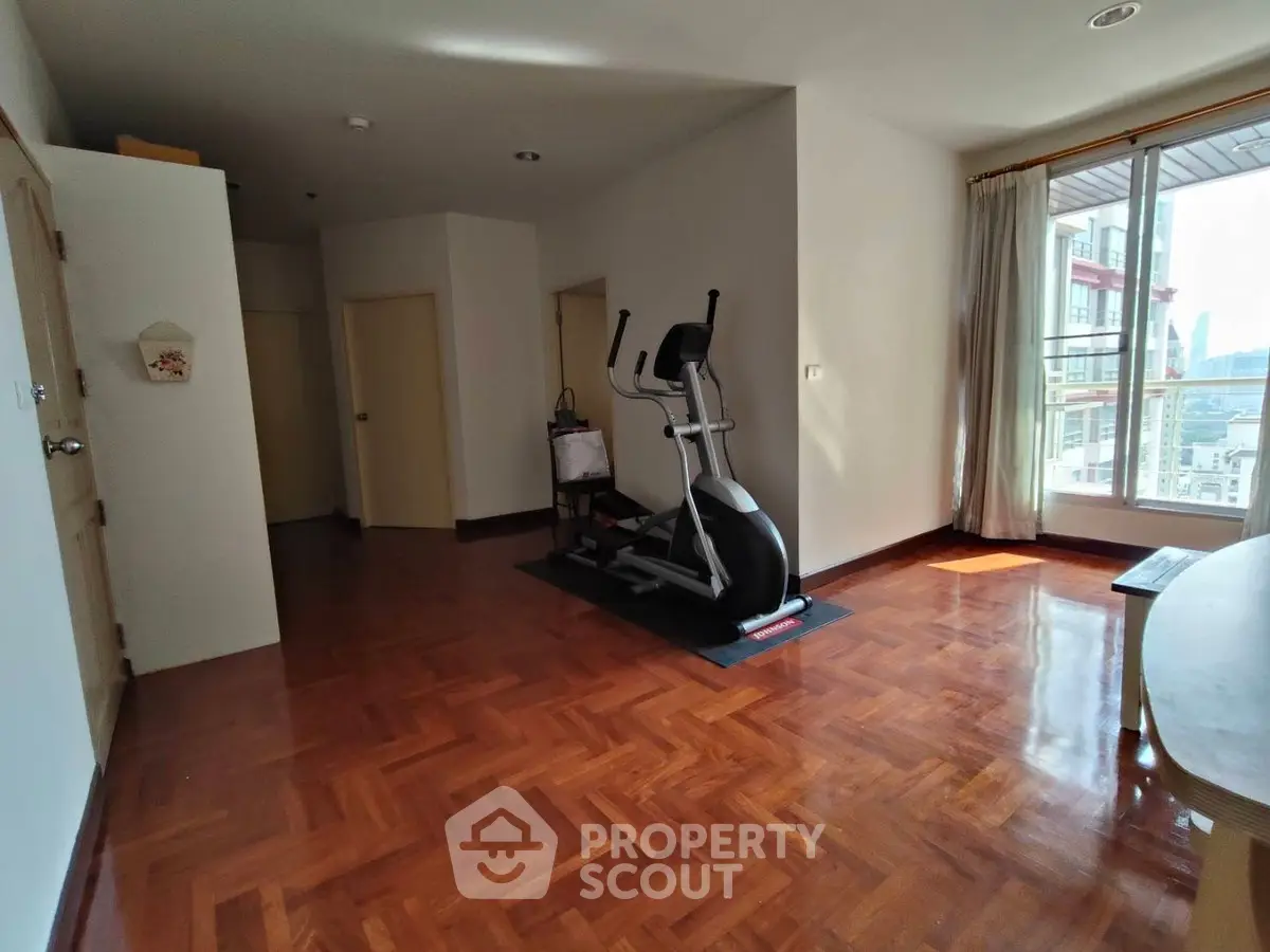 Spacious room with exercise equipment and wooden flooring, large window with city view.