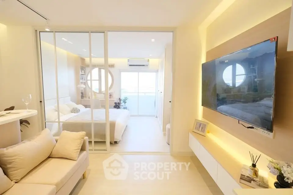 Modern open-plan living room with cozy sofa and wall-mounted TV, leading to a bright bedroom.
