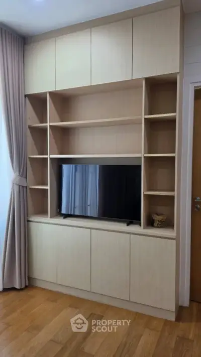 Modern living room with built-in wooden shelves and TV unit, elegant curtains, and wooden flooring.