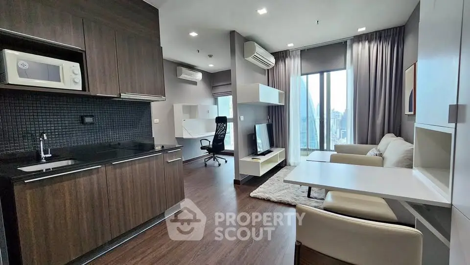 Modern open-plan living room with kitchen, featuring sleek dark wood cabinets and large windows.