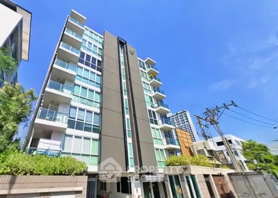 Modern multi-story apartment building with balconies and lush greenery under a clear blue sky.