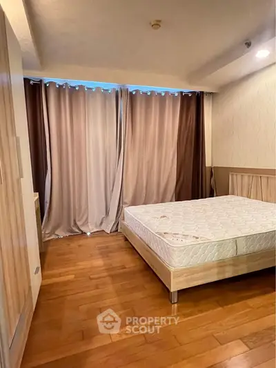 Spacious bedroom with wooden flooring and large window draped with elegant curtains.