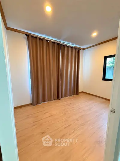 Spacious empty room with wooden flooring and large window with curtains.