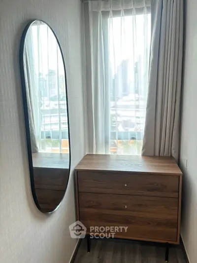 Stylish bedroom corner with modern dresser and oval mirror, city view through window.