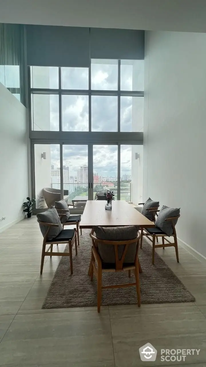 Spacious living room with high ceiling and large windows offering panoramic city views, complemented by a stylish dining set and cozy seating area.