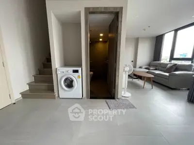 Modern living room with washing machine and staircase, featuring sleek furniture and large windows.