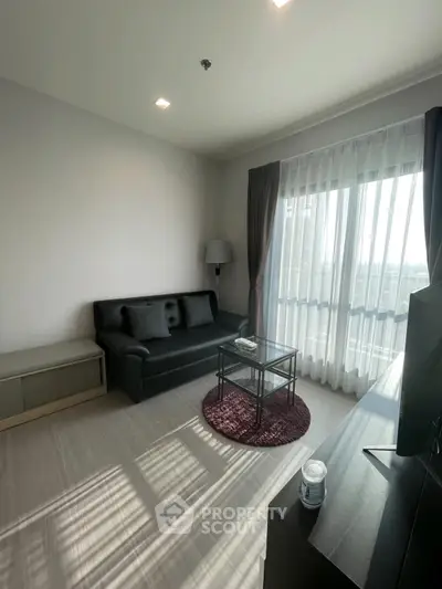 Modern living room with sleek black sofa, glass coffee table, and large window offering natural light.