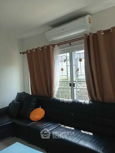 Cozy living room with black leather sofa and air conditioning, featuring large windows with brown curtains.
