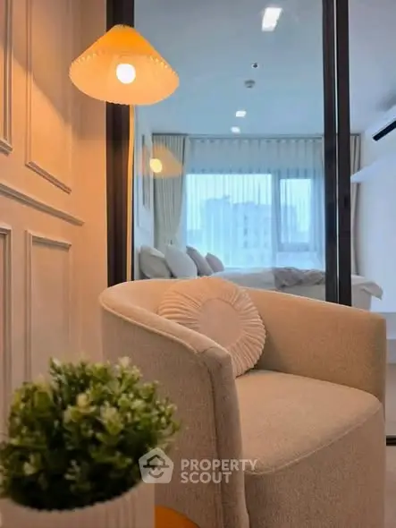 Cozy modern living room with elegant chair and soft lighting, overlooking a serene bedroom through glass doors.