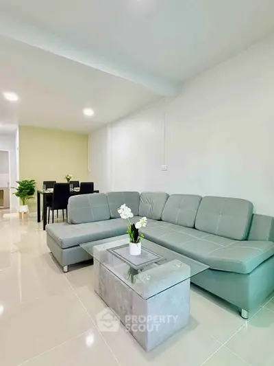 Modern living room with sleek gray sofa and glass coffee table, open layout leading to dining area.
