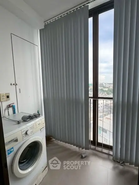 Compact laundry area with washing machine and city view through vertical blinds.