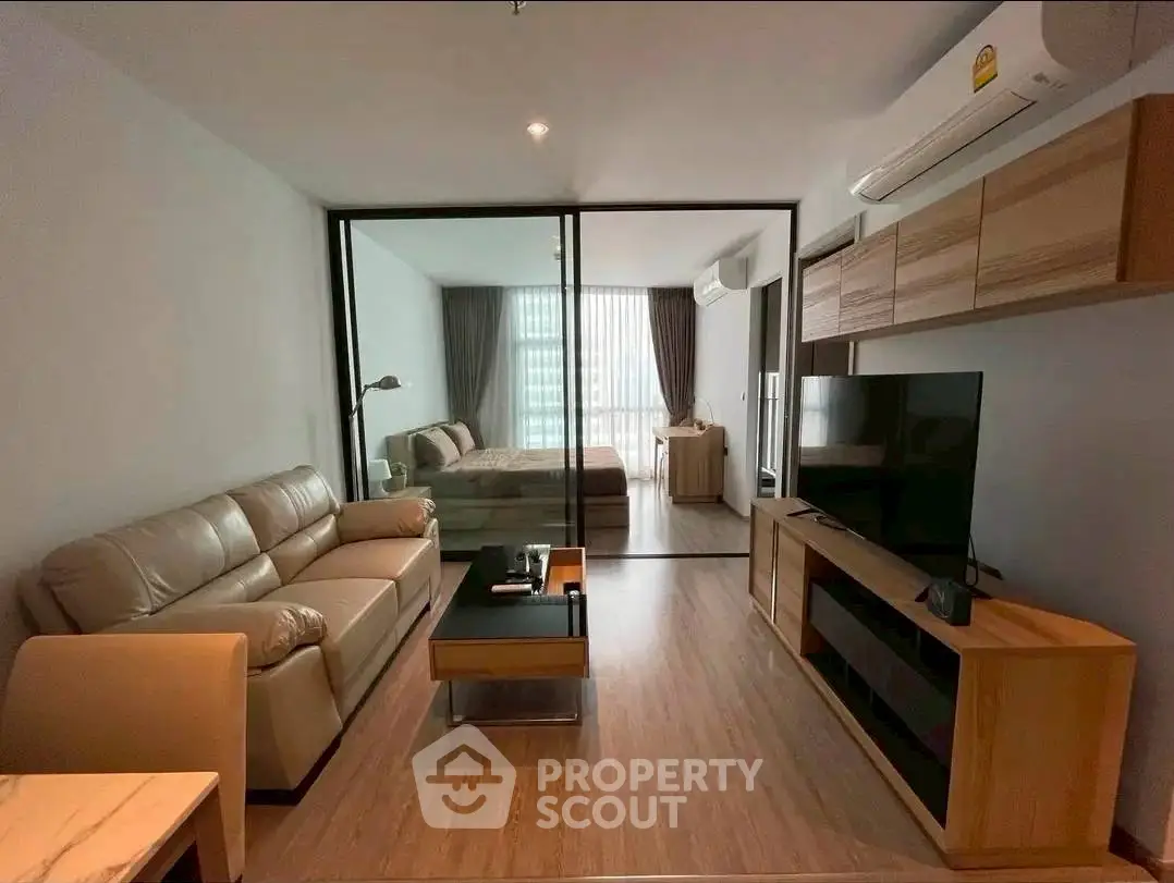 Modern living room with glass partition leading to bedroom, featuring sleek furniture and natural light.