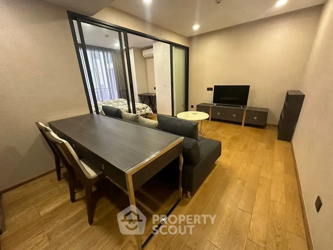 Modern living room with wooden flooring, sleek furniture, and a view into the bedroom through glass doors.