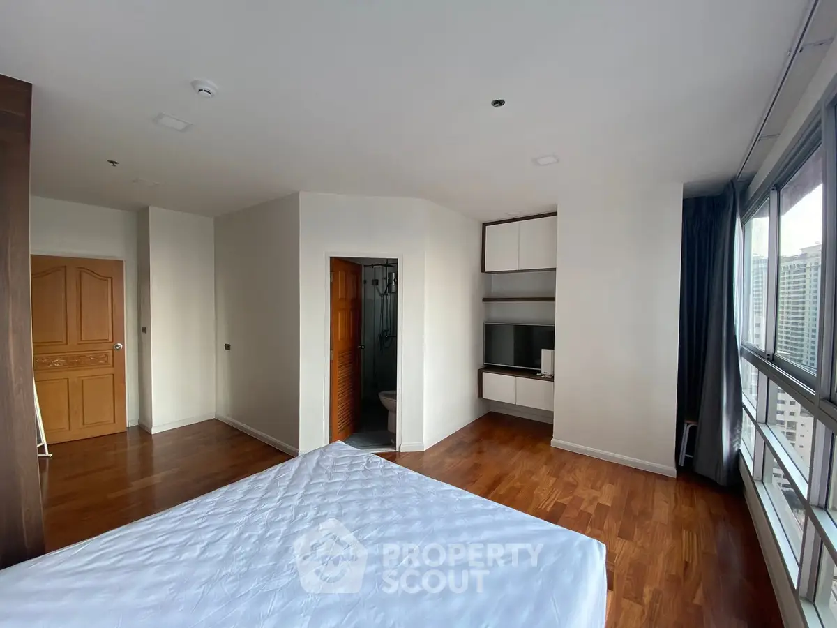 Spacious bedroom with hardwood floors and city view, featuring built-in shelves and a cozy ambiance.