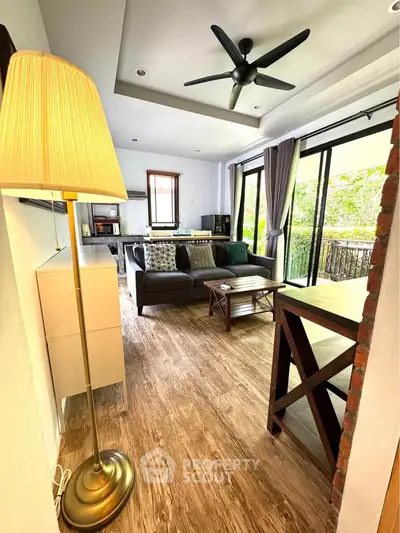 Charming living room with wooden flooring, cozy seating, and large windows offering lush garden views.