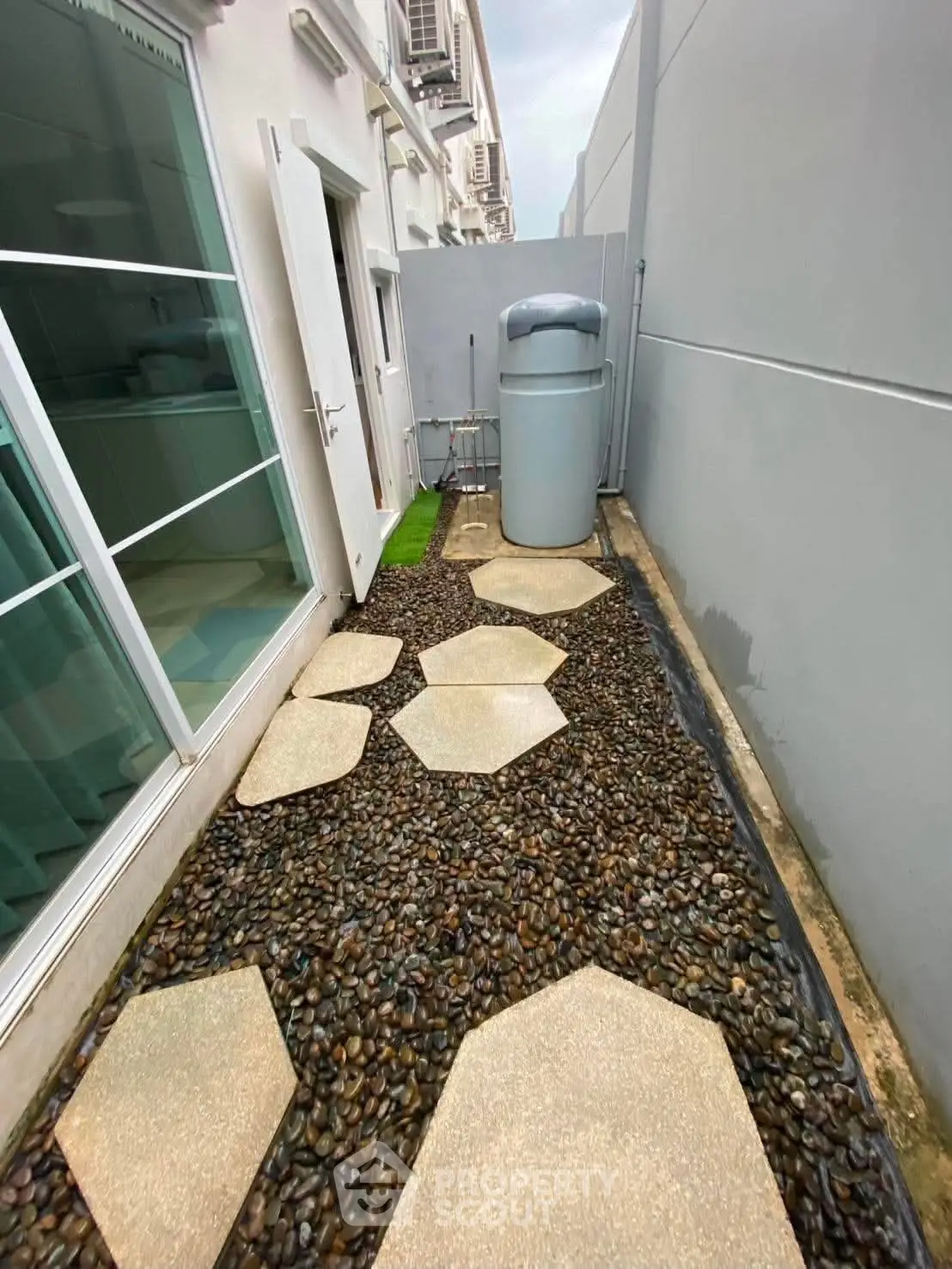 Modern exterior walkway with hexagonal stepping stones and pebble design, featuring sleek water tank and glass windows.