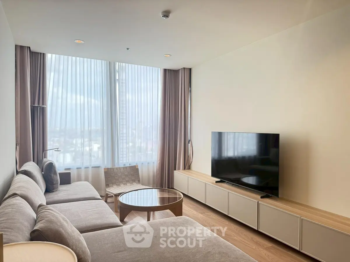 Modern living room with large sofa, flat-screen TV, and city view through floor-to-ceiling windows.