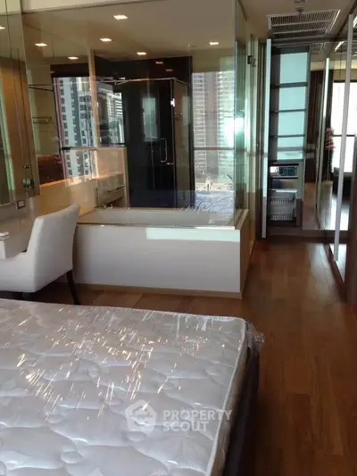 Luxurious bedroom with glass-walled bathtub and city view, featuring modern furnishings and hardwood floors.