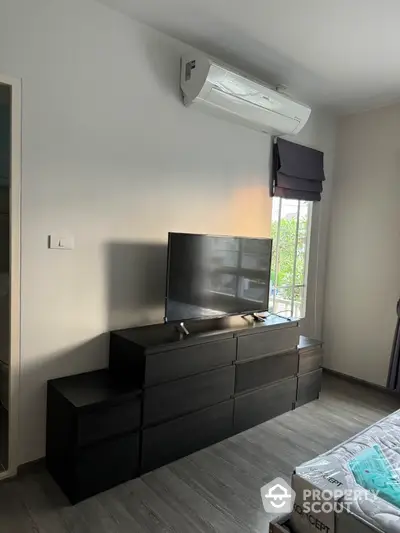 Modern bedroom with sleek black dresser and mounted TV, featuring air conditioning and natural light.