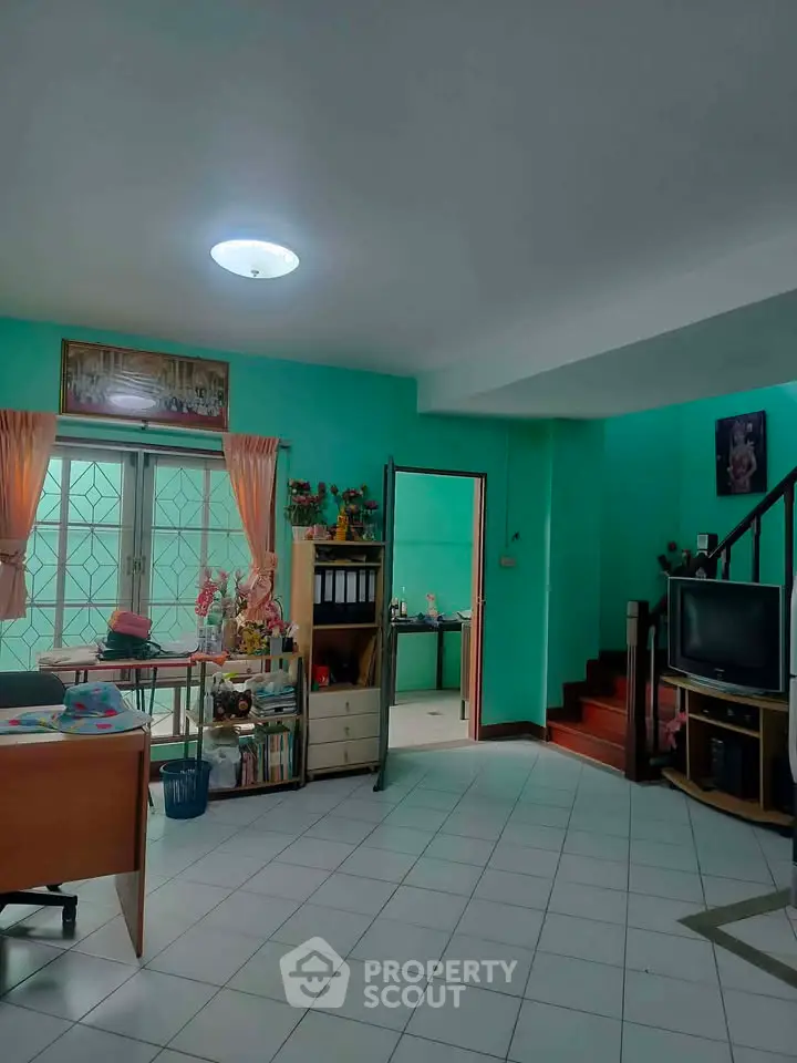 Spacious living room with vibrant teal walls and natural light, featuring a cozy workspace and TV area.