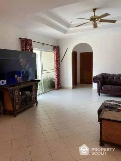 Spacious living room with elegant sofa and large TV, featuring natural light and ceiling fan.