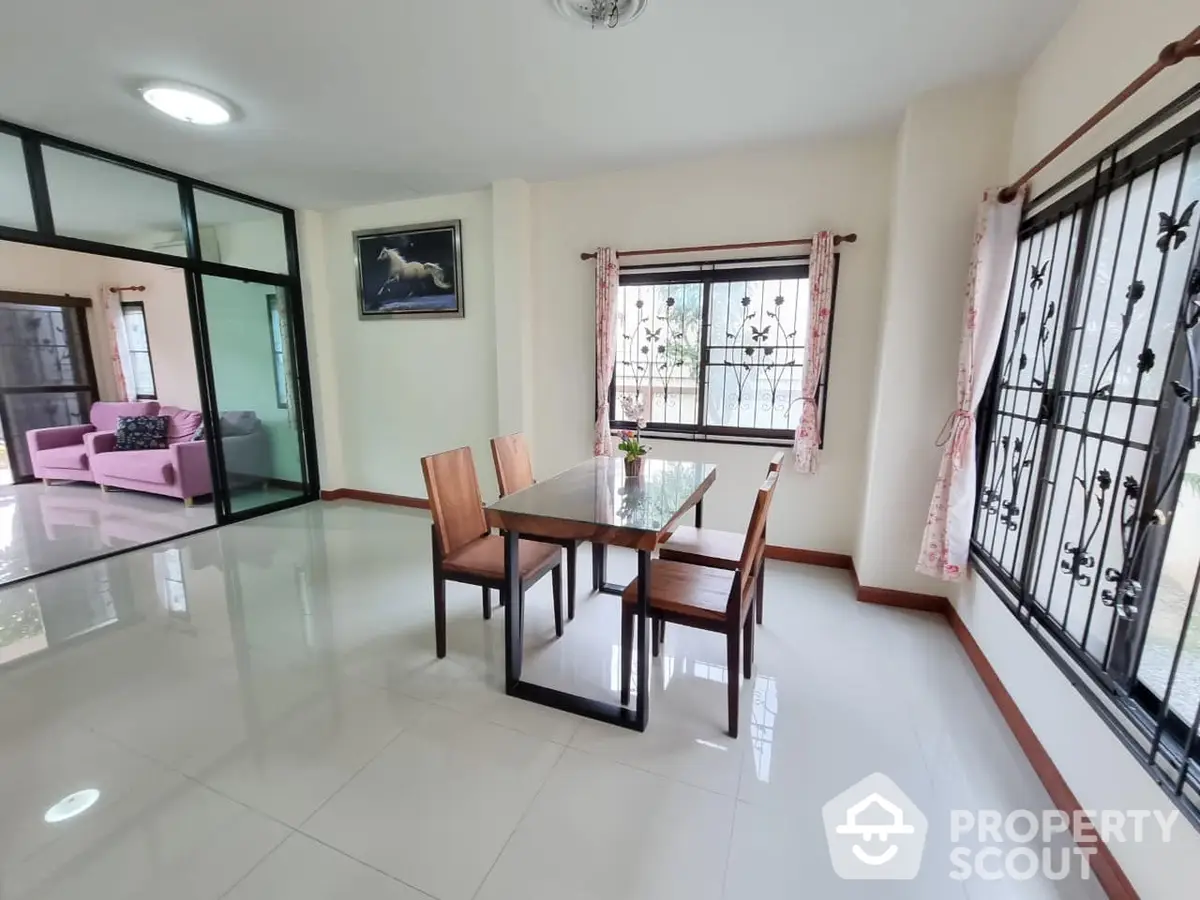 Spacious and well-lit living room with glossy tiled floors, large windows with protective grills, and a sleek dining table set, leading to a cozy pink-themed seating area.