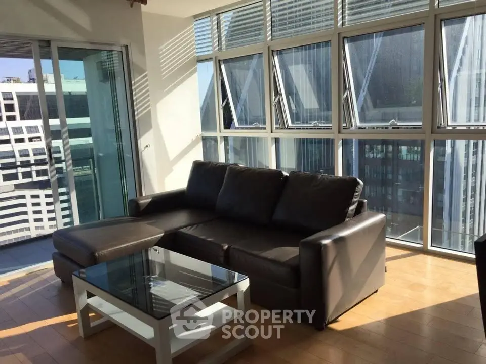 Modern living room with large windows and city view, featuring a sleek black sofa and glass coffee table.