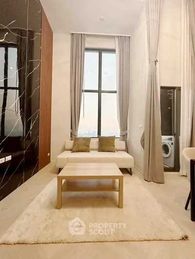 Modern living room with high ceilings and large window, featuring a cozy sofa and washing machine.