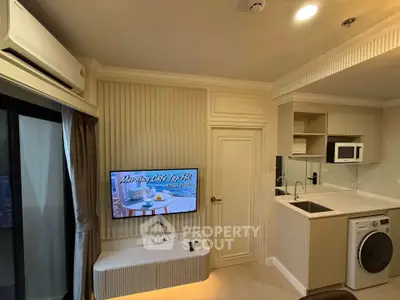 Modern living room with TV, air conditioning, and compact kitchen featuring washing machine and microwave.