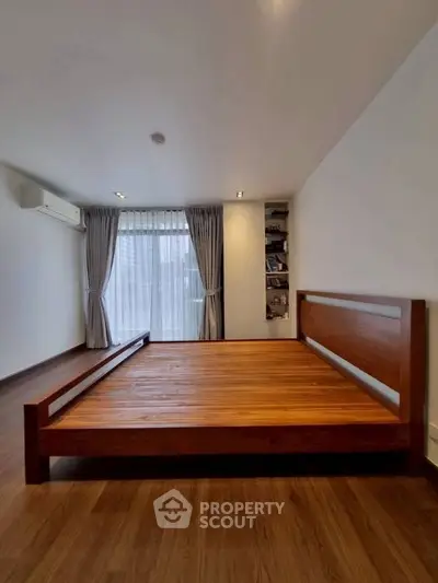 Spacious bedroom with wooden flooring and large window, featuring a minimalist wooden bed frame.