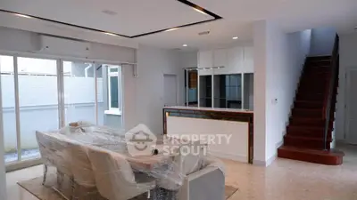 Modern open-plan living room with sleek kitchen and staircase, featuring large windows and elegant dining area.