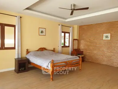 Spacious bedroom with wooden bed and brick accent wall, featuring natural light and ceiling fan.