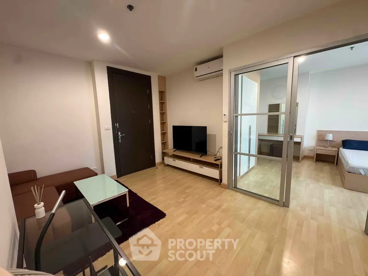 Modern living room with wooden flooring, glass coffee table, and TV unit, adjacent to a cozy bedroom.