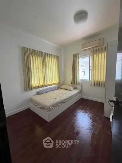 Cozy bedroom with wooden flooring and natural light from large windows, featuring a comfortable bed and air conditioning.
