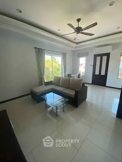 Spacious living room with modern sofa and glass coffee table, featuring large windows and ceiling fan.