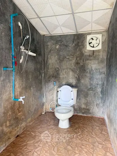 Rustic bathroom with exposed plumbing and ceiling fan, featuring a simple toilet setup.