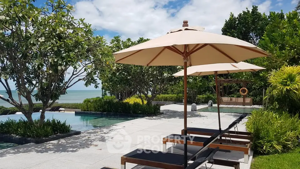 Luxurious beachfront pool area with sun loungers and umbrellas, surrounded by lush greenery and ocean views.