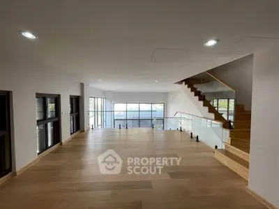 Spacious modern living room with wooden flooring and glass staircase, offering ample natural light.