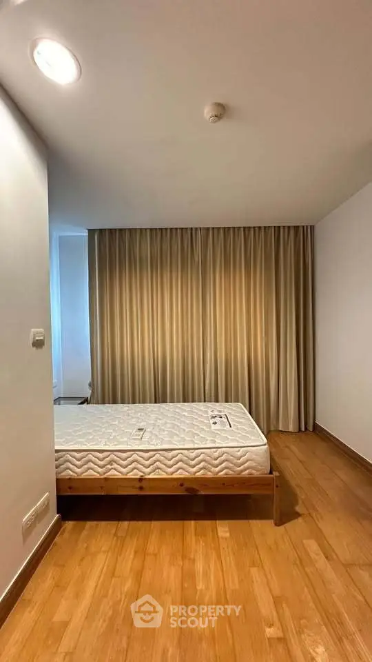 Spacious bedroom with wooden flooring and large window draped with curtains, featuring a comfortable mattress.